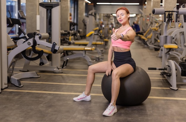 Roodharige, vrolijke fitte vrouw zit op een fitball in een moderne sportschool Toont duim omhoog