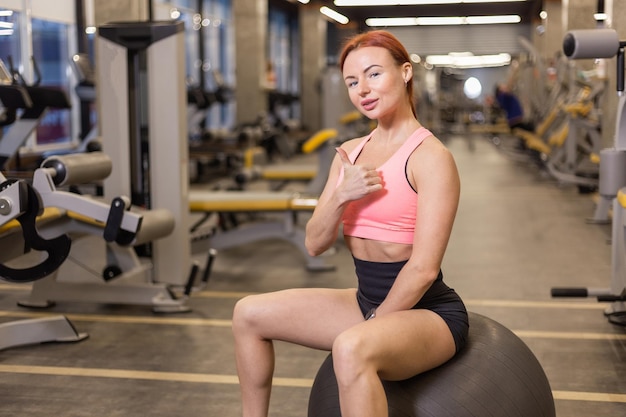 Roodharige, vrolijke fitte vrouw zit op een fitball in een moderne sportschool Toont duim omhoog