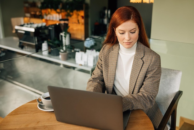 Roodharige vrouw die op laptop typt en mobiele telefoon vasthoudt terwijl ze in een cafex9xA zit