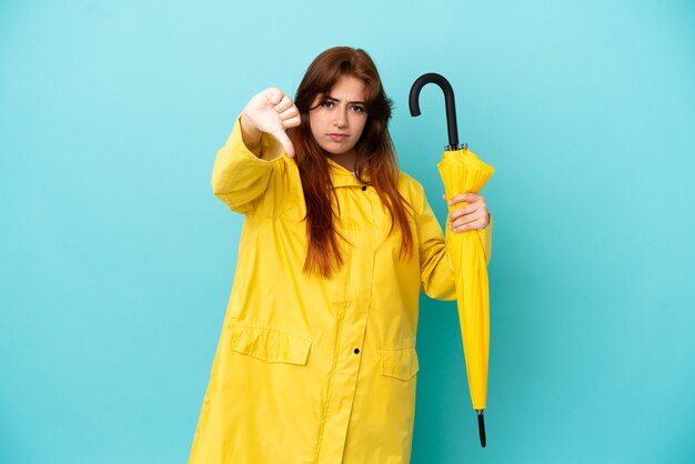 Roodharige vrouw met een paraplu geïsoleerd op een blauwe achtergrond met duim omlaag met negatieve uitdrukking