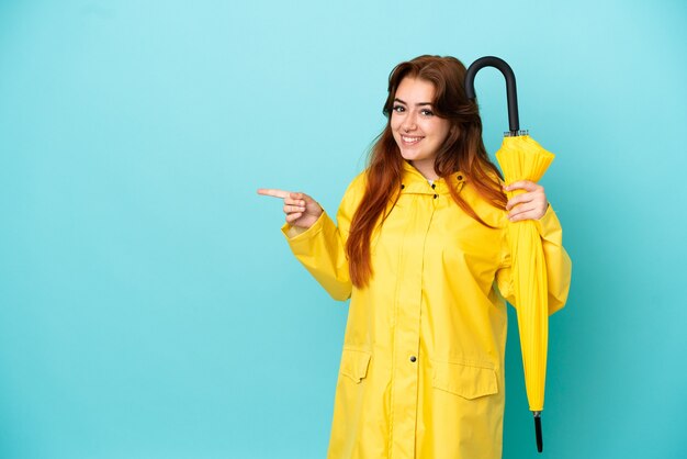 Roodharige vrouw met een paraplu geïsoleerd op een blauwe achtergrond wijzende vinger naar de zijkant