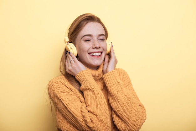 Roodharige vrouw met sproeten luistert naar muziek op een koptelefoon die lacht op een gele muur