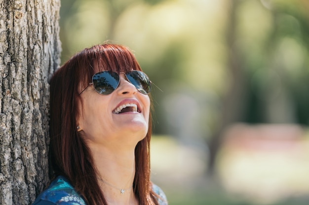 Roodharige vrouw met zonnebril lachend leunend tegen een boom op een zonnige dag