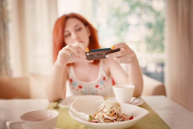 Roodharige vrouw neemt foto's van salade op smartphone camera witte keramische gerechten op tafel salade van ch...