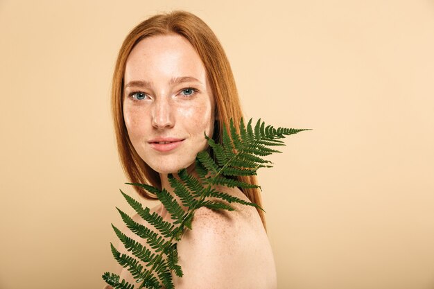 Roodharige vrouw stond geïsoleerd over beige muur met blad bloem plant.