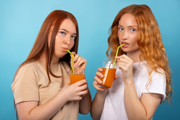 Roodharige vrouwen drinken sinaasappelsap uit gele rietjes op een blauwe muur