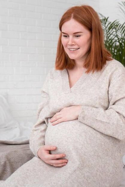 Roodharige zwangere vrouw in warme, knusse jurk legt handen op de buik terwijl ze thuis in de slaapkamer zit