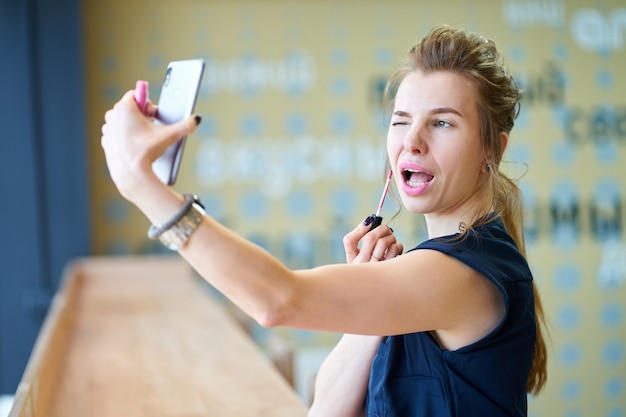 Roodharigemeisje die lippenstift zelf aanzetten, die bij bezinning op smartphone letten op