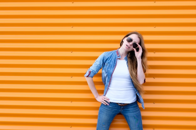 Roodharigemeisje in jeansoverhemd met zonnebril dichtbij oranje muur