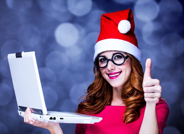 Roodharigemeisje in kerstmishoed en glazen met laptopcomputer op grijs