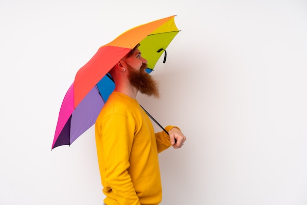 Roodharigemens die met lange baard een paraplu over geïsoleerde witte muur houden kijkend aan de kant