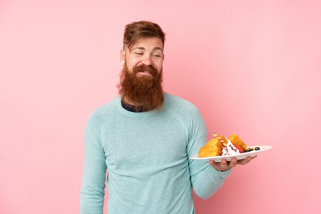 Roodharigemens met de lange wafels van de baardholding over geïsoleerde roze muur met gelukkige uitdrukking