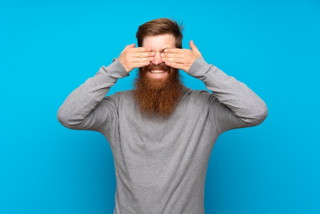 Roodharigemens met lange baard over blauwe muur die ogen behandelen door handen