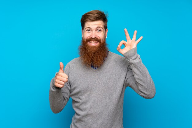 Roodharigemens met lange baard over geïsoleerd blauw dat ok teken en duim op gebaar toont