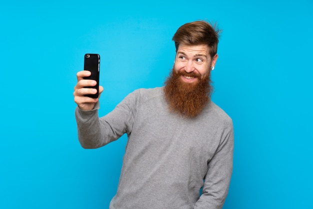 Roodharigemens met lange baard over geïsoleerd blauw die een selfie maken