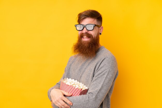 Roodharigemens met lange baard over geïsoleerde gele muur met 3d glazen en holding een grote emmer popcorns