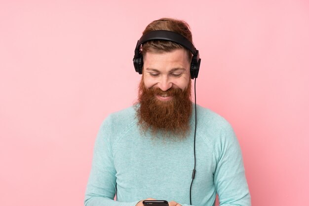 Roodharigemens met lange baard over geïsoleerde roze het luisteren muziek en het kijken aan mobiel