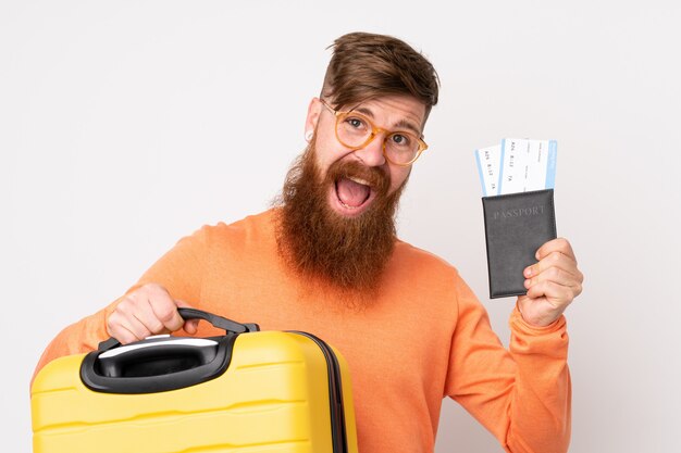 Roodharigemens met lange baard over geïsoleerde witte muur in vakantie met koffer en paspoort