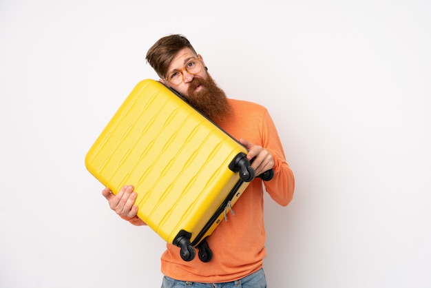 Roodharigemens met lange baard over geïsoleerde witte muur in vakantie met reiskoffer en ongelukkig