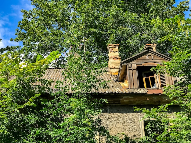 Roof of dilapidated clapboard house with an attic window and greenery around house