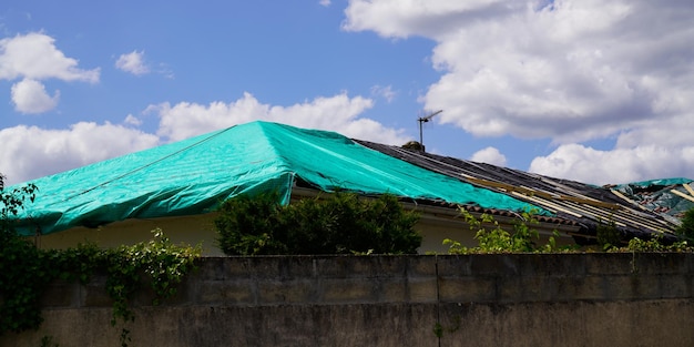 Roof house temporary repair on badly storm damaged roof with a big leaky hole in shingles and rooftop covered hole with green plastic tarp