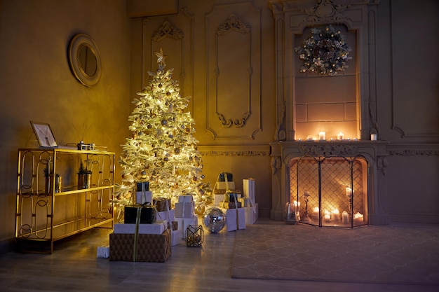 Photo room decorated to christmas celebration studio. white christmas tree with white and gold balls in a dark room with a decorative fireplace and candles among boxes with gifts