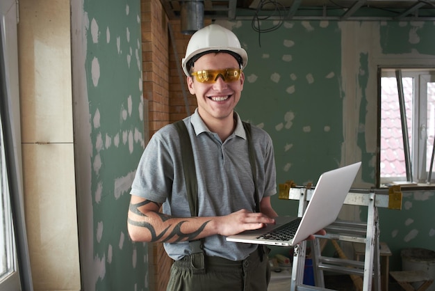 Room repair. Builder in helmet and glasses stands with a laptop in his hands on the background of the construction