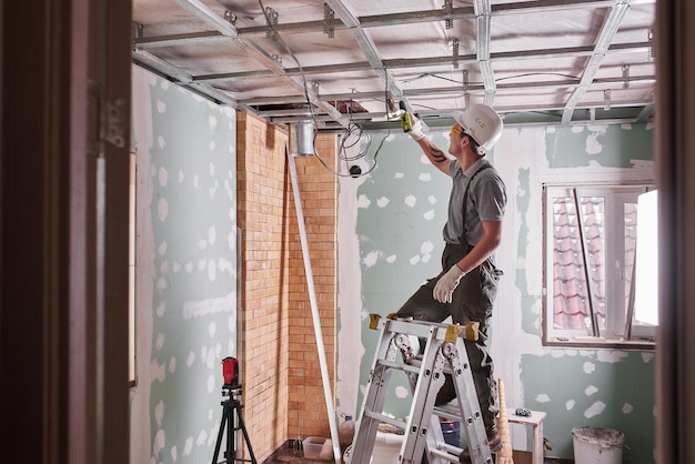 Riparazione della stanza. finitura interna. il giovane costruttore realizza un soffitto in cartongesso, in piedi su una scala a pioli