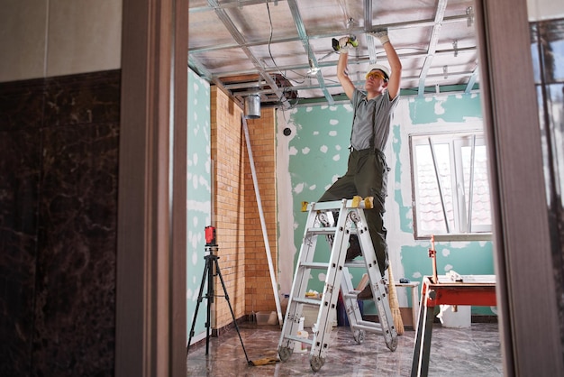 Foto riparazione della stanza. finitura interna. il giovane costruttore realizza un soffitto in cartongesso, in piedi su una scala a pioli