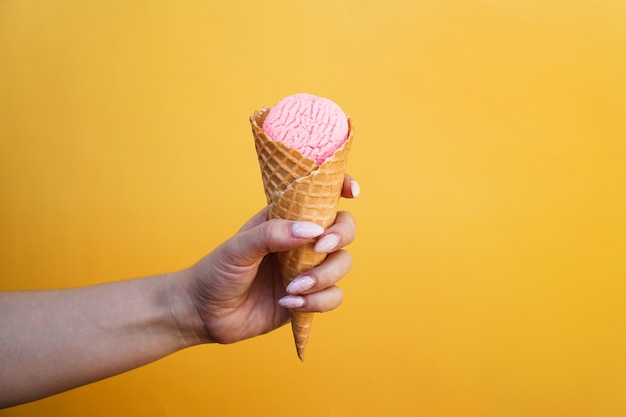 Roomijs in wafelkop in vrouwelijke hand op gele achtergrond. Roze aardbeienroomijs in een wafelhoorntje