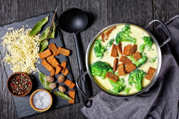 Roomkaassoep met broccoli en roggecroutons in een pot op een donkere houten tafel met lepel en ingrediënten, horizontale weergave van bovenaf, plat gelegd