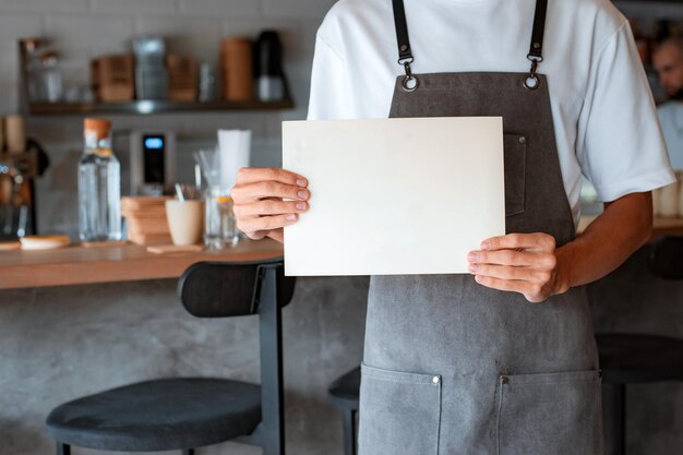 Ropped photo of unknown man coffee shop owner or cafe worker holding blank sheet in hands while standing against background of wooden counter in coffeehouse, mockup for advertising. Small business