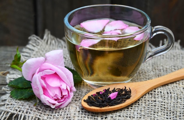 Rose herbal tea in a glass cup with pink petals and fresh flower on a rustic wooden table