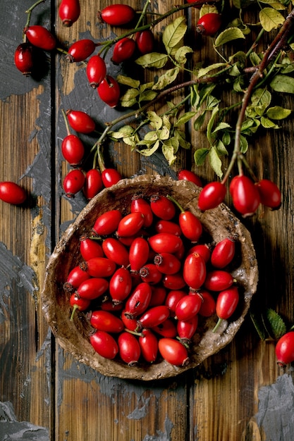 Rose hip berries on ceramic plate with branch and leaves on old wooden plank background. Autumn reserves of vitamins for the winter. Flat lay, close up