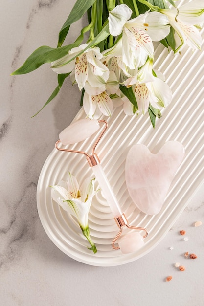 A rose quartz cosmetic roller massager a gua sha scraper and astromeria flowers lie on a white podium in the form of an arch Top view selfcare