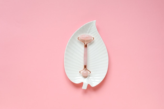 Rose quartz facial massage roller on white leaf shaped plate on pink background.