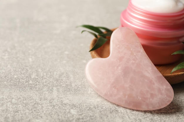 Rose quartz gua sha tool and jar of cream on grey table closeup Space for text