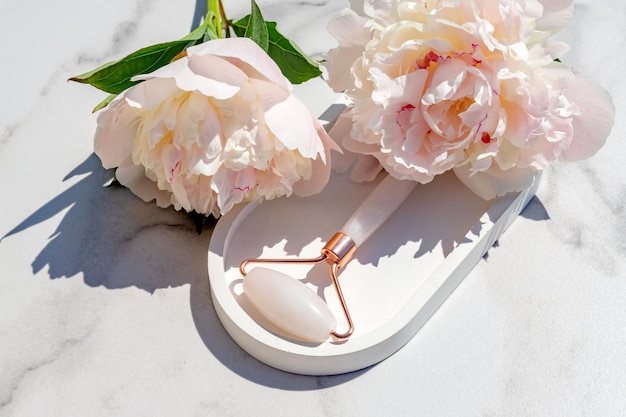 Rose Quartz jade roller on concrete tray on marble background with peony flowers Anti age lifting and toning treatment massaging at home Copy space