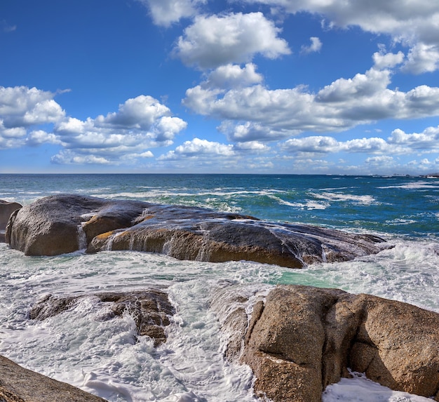 Rotsen in de oceaan onder een blauwe bewolkte hemel met kopieerruimte Schilderachtig landschap van strandgolven die over rotsblokken of grote stenen in de zee wassen op een populaire zomerlocatie in Kaapstad, Zuid-Afrika