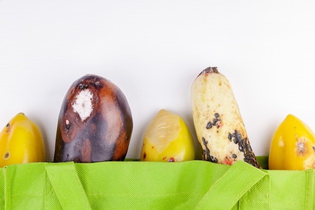 Rotten organic vegetables in green reusable shopping bag on white background