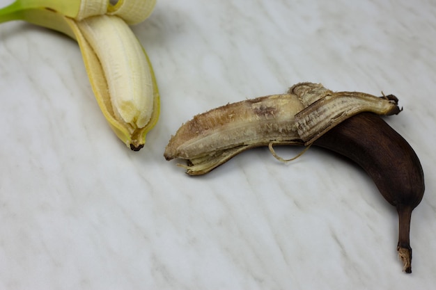 Rotten spoiled banana in open peel next to fresh banana fruit on white table