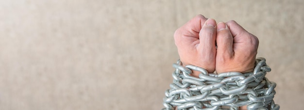 the rough hands of a worker a white male are wrapped in a chain as a concept of slavery or imprisonment closeup photo of the hands of a slave with a chain on a gray background with copy space