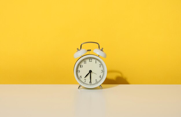 A round alarm clock stands on a white table, half past seven in the morning. Yellow background