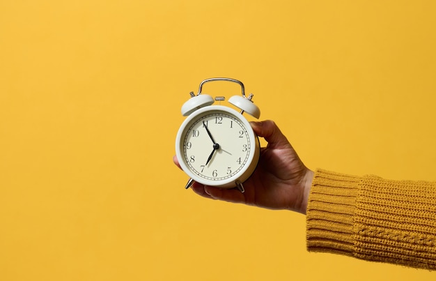 Round alarm clock in a woman's hand, the time is five minutes to seven in the morning. Yellow background