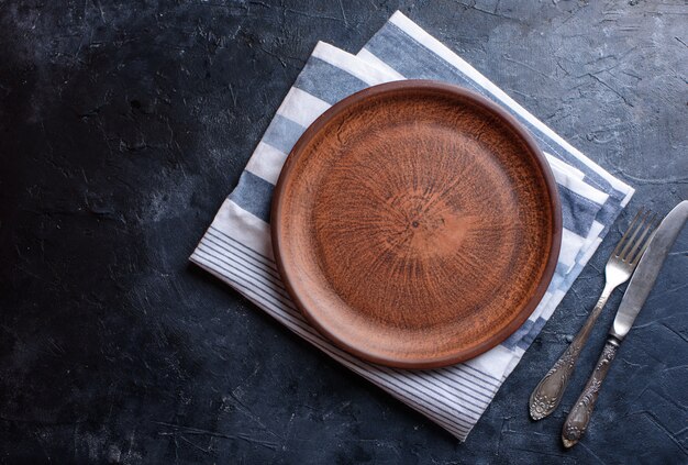 Round brown plate, for food, fork, knife, towel, on a dark background. Flat lay