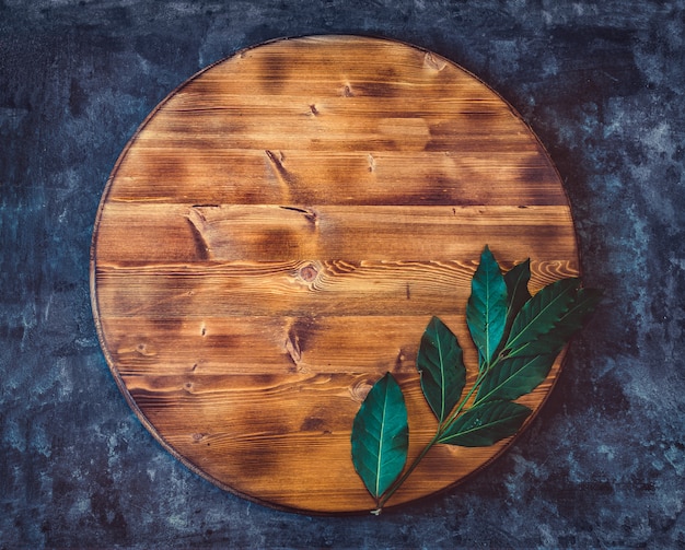 Round empty wooden cutting board with bay leaf branch on a dark gray textured background. Top view. Copy space