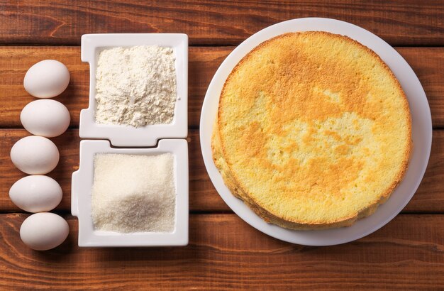 Round homemade golden biscuit cake on a plate, flour, sugar and eggs on wooden table. Ingredients for the cake. The cake and the ingredients from which it was prepared