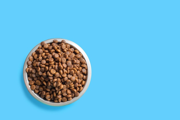 Round metal bowl full of crunchy dry food for dogs or cats on blue background, top view, copy space. Pet care concept. Delicious and healthy food for domestic cats and dogs. Diet food for pets.