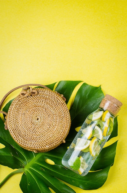 Round rattan bag and glass bottle with lemonade on a Monstera leaf. 