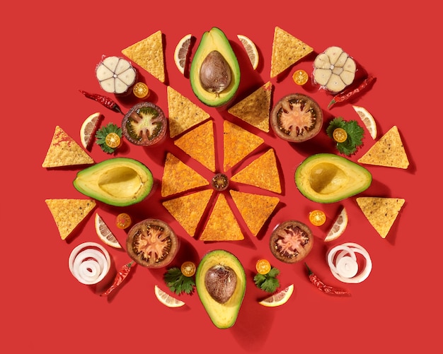 Round traditional mexican pattern with corn nachos chips, fresh natural fruits, vegetables, spices, chilli greens - ingredients for Guacamole dressing on a red background. Flat lay.